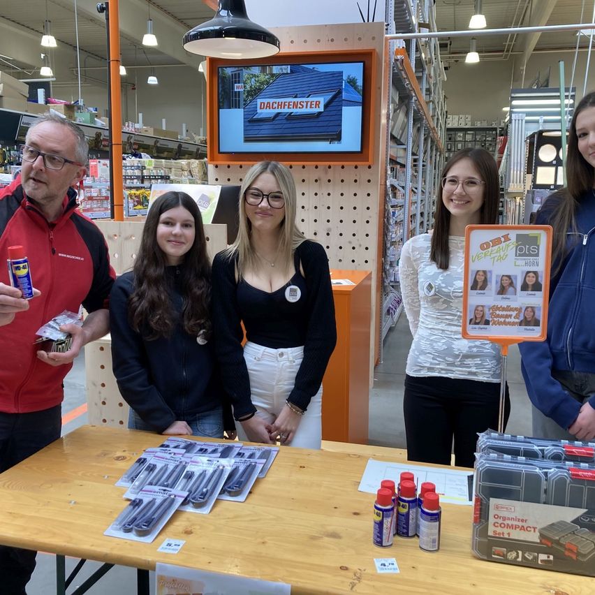 Fünf Personen stehen in einem Geschäft und lächeln. Ein Tisch hat verschiedene Werkzeuge und Produkte. Eine Frau hält ein Schild mit Fotos. Dahinter zeigt ein Monitor 'DACHFEHNSTER' an.
