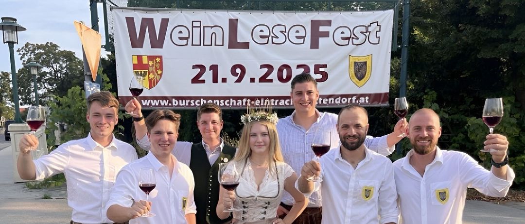 Eine Gruppe junger Erwachsener in traditioneller bayerischer Kleidung, mit Weinglas in der Hand, posiert für ein Foto vor einem Banner für das Wein Lese Fest 2025.