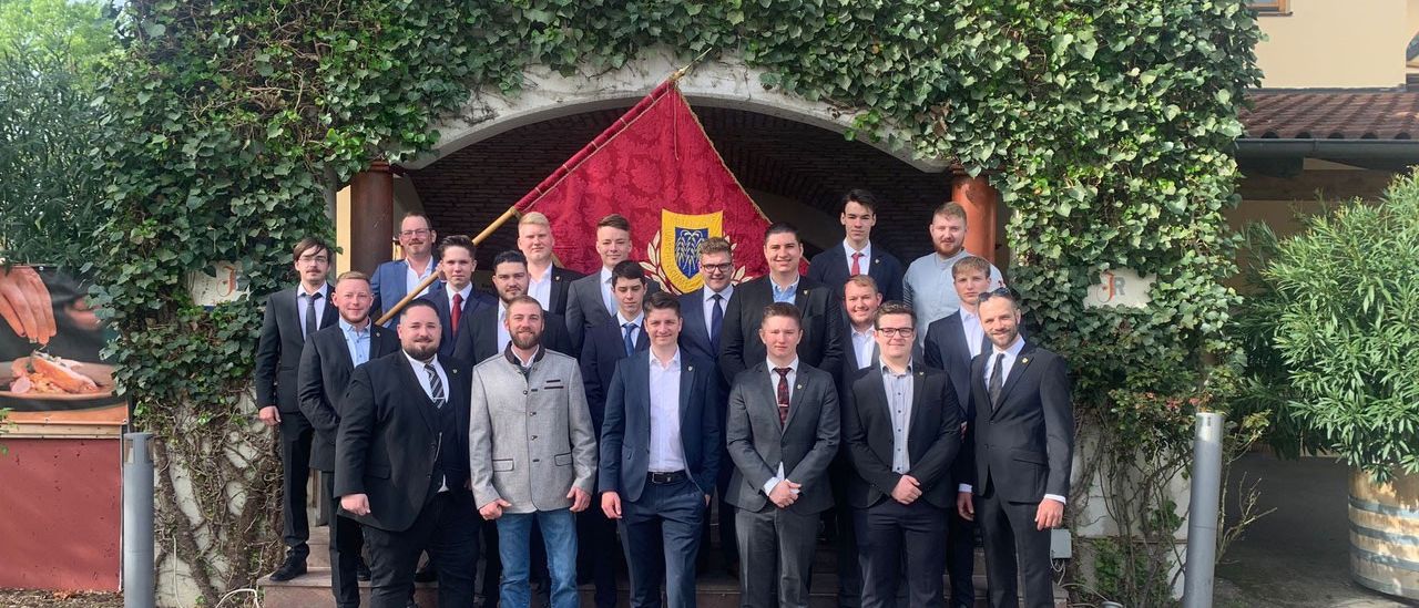 Eine Gruppe von formell gekleideten Männern steht auf den Stufen eines Gebäudes, lächelt und posiert für ein Foto. Hinter ihnen befindet sich eine Ziegelmauer mit einer roten Flagge mit einem goldenen Logo.