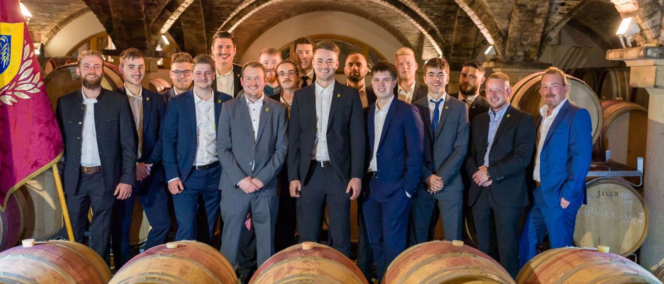 Eine Gruppe von Männern in Anzügen und Krawatten steht hinter Fässern in einem Keller. Sie lächeln und scheinen für ein Foto zu posieren. Die Fässer sind aus Holz und haben Etiketten darauf.