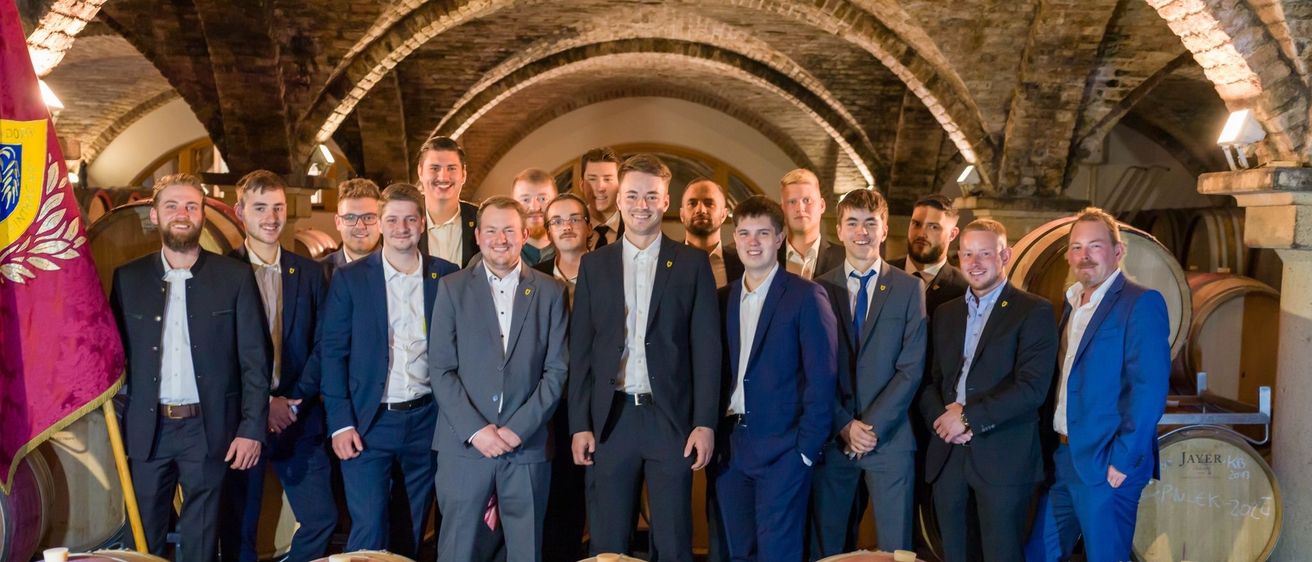 Eine Gruppe formell gekleideter Männer steht in einem Weinkeller und posiert für ein Foto vor Holzfässern.