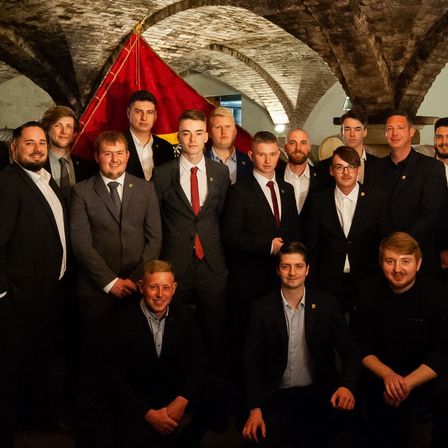 Eine Gruppe von Männern in Anzügen und Krawatten posiert für ein Foto in einem gewölbten, schwach beleuchteten Raum mit einer roten Fahne, die von der Decke hängt.