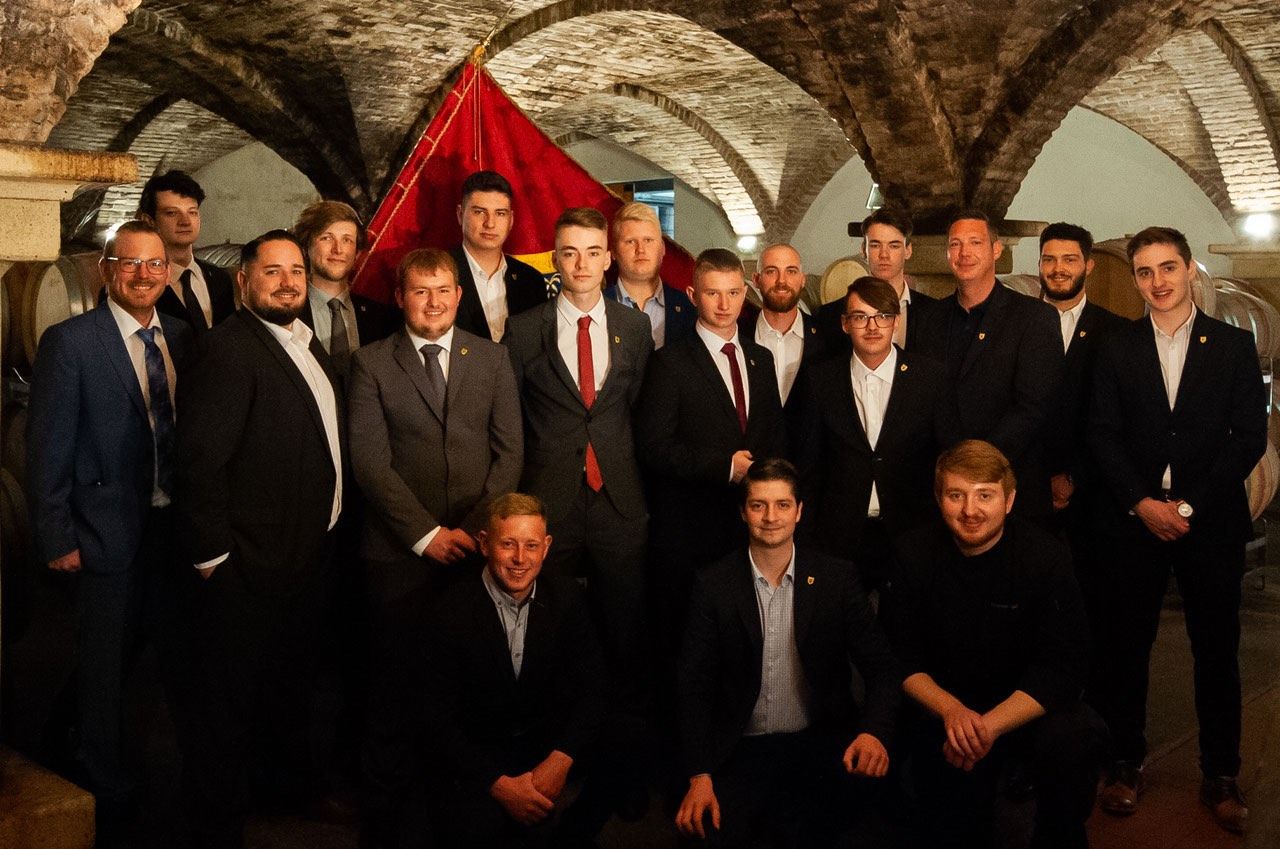 Eine Gruppe von Männern in Anzügen und Krawatten posiert für ein Foto in einem gewölbten, schwach beleuchteten Raum mit einer roten Fahne, die von der Decke hängt.