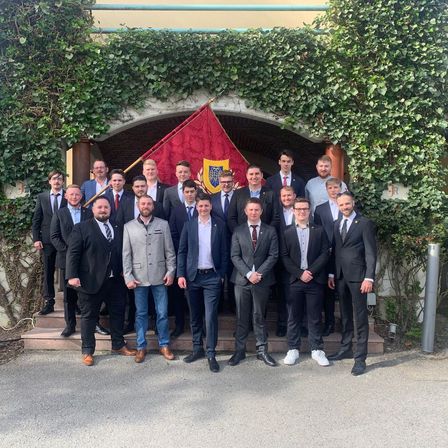 Eine Gruppe von Männern in Anzügen steht vor einem Bogen mit Efeu. Sie posieren für ein Foto, wobei einige Krawatten und andere eine Brille tragen.