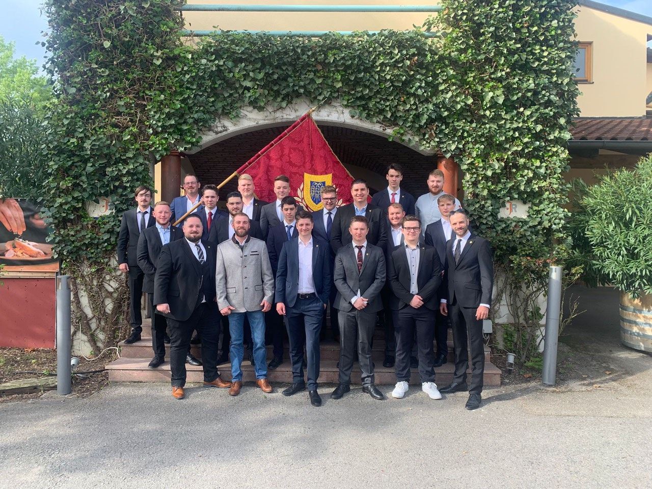 Eine Gruppe von Männern in Anzügen steht vor einem Bogen mit Efeu. Sie posieren für ein Foto, wobei einige Krawatten und andere eine Brille tragen.