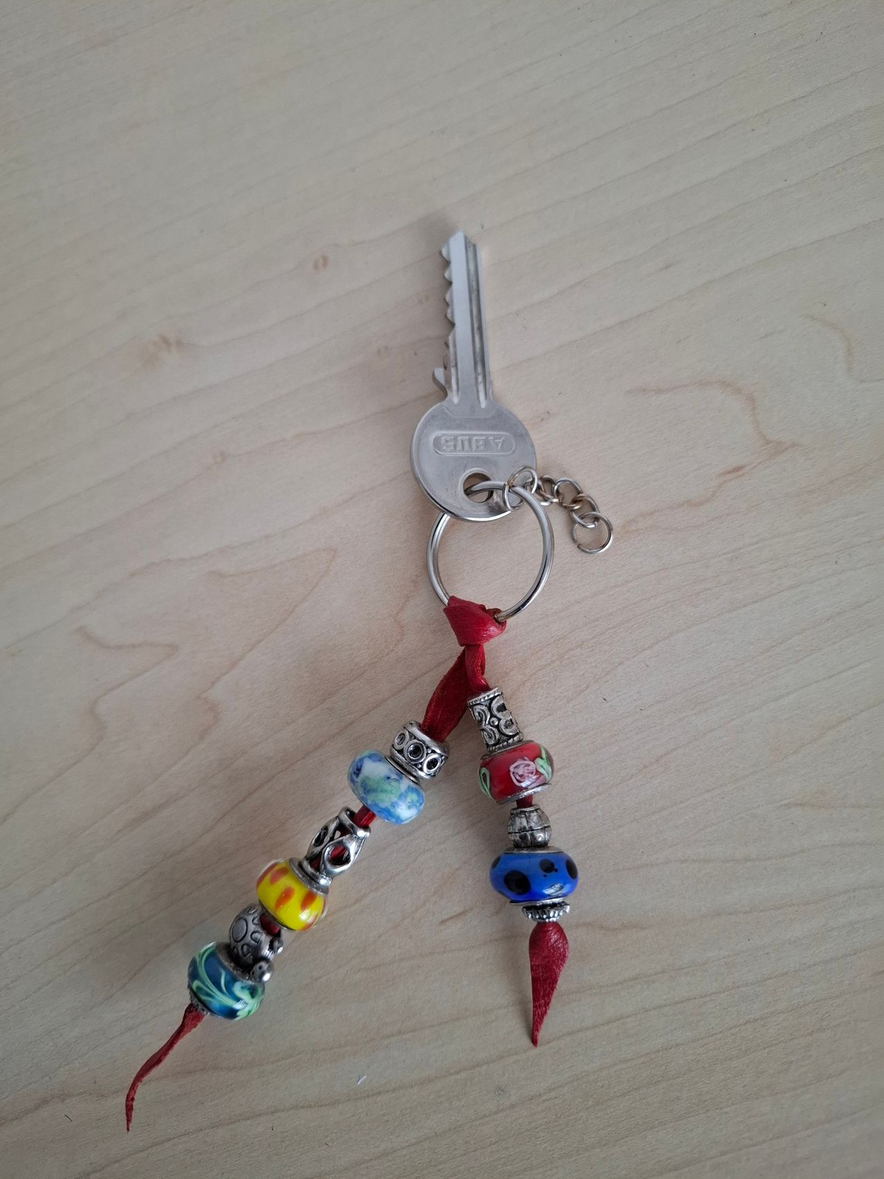 Ein silberner Schlüsselanhänger mit einem roten Band und bunten Perlen liegt auf einer Holzoberfläche. Der Schlüsselanhänger enthält einen silbernen Schlüssel.