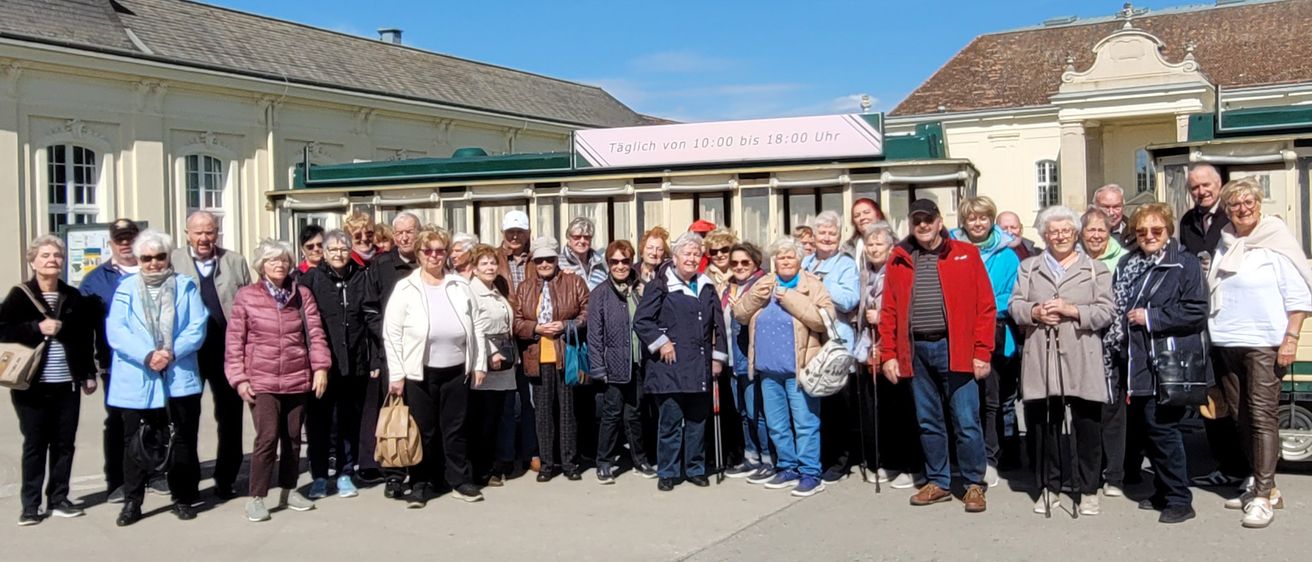 Eine Gruppe von Menschen posiert für ein Foto vor einem Gebäude. Das Gebäude hat ein Schild mit der Aufschrift 'Taglich von 10:00 bis 18:00 Uhr.' Die Menschen tragen Winterkleidung.
