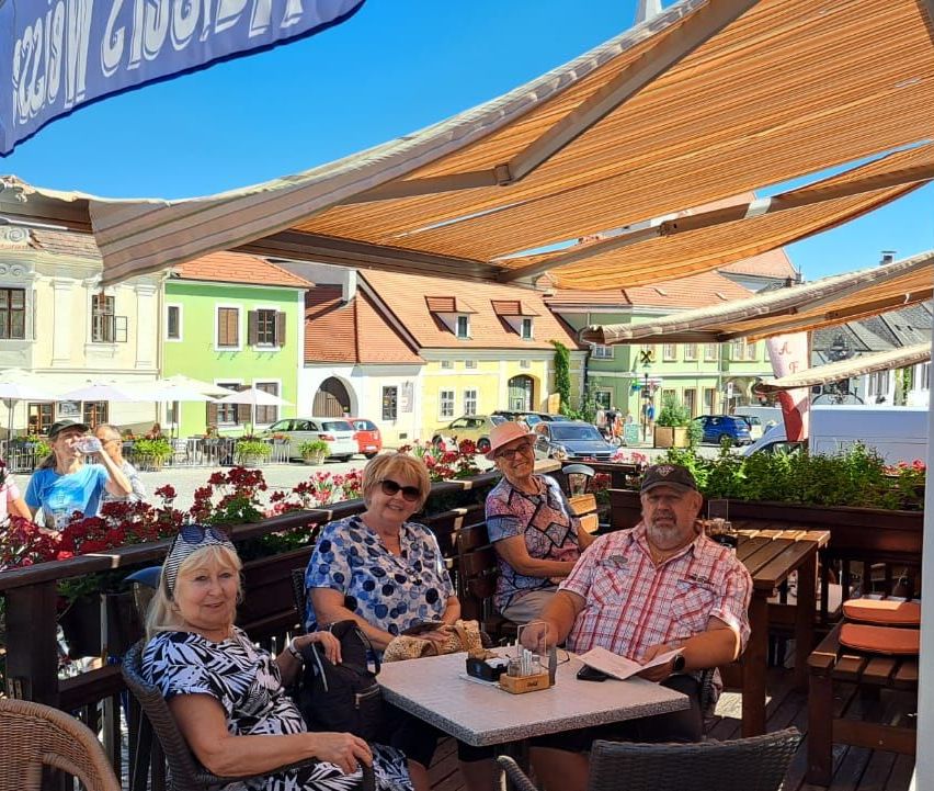 Four people are sitting outside at a restaurant. Two women and two men. One man is reading a book. The other man is talking to the women. There are flowers and buildings in the background.