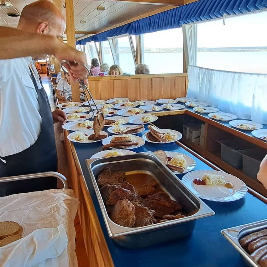 A man in a white shirt and apron serves food from a buffet on a boat. Plates of food are arranged on the table.