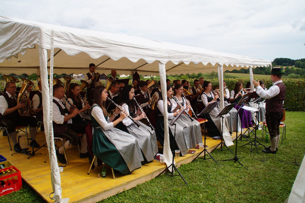 Eine Gruppe von Musikern, einige sitzend und andere stehend, spielen unter einem weißen Zelt. Sie spielen verschiedene Instrumente, darunter Klarinetten und Trompeten. Die Bühne ist mit Gras bedeckt.