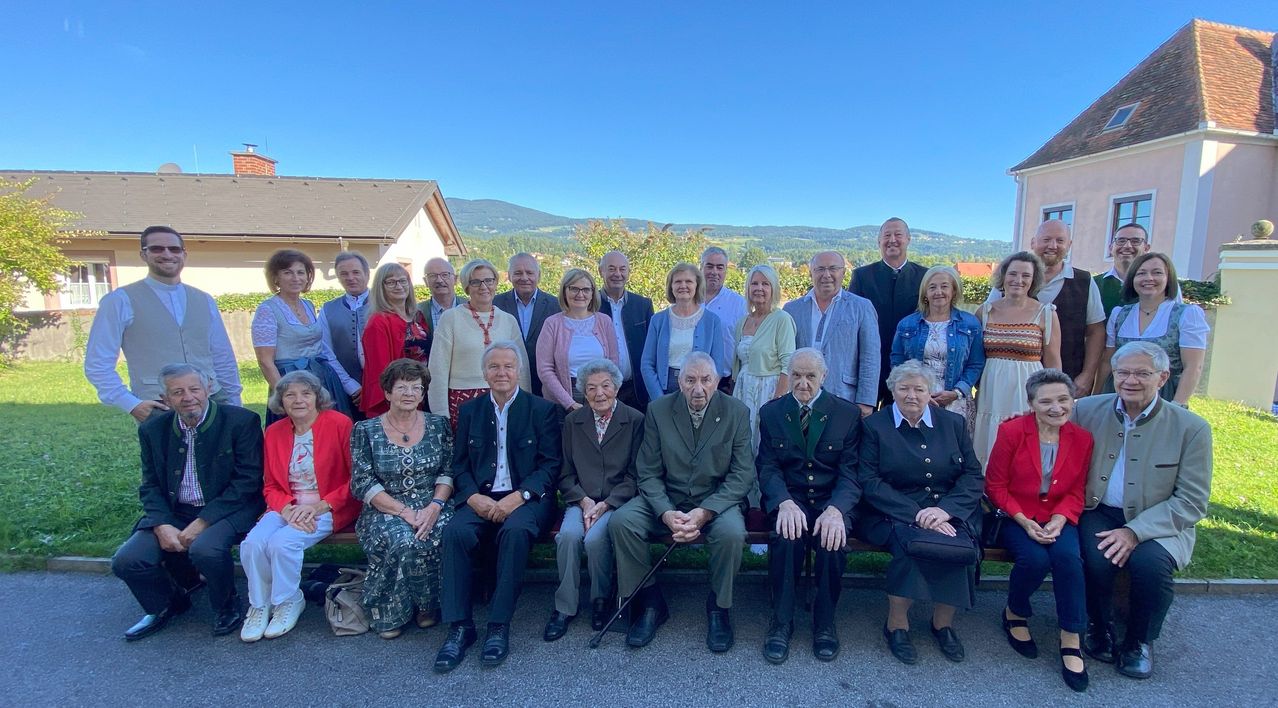 Eine Gruppe von Menschen, einige in formeller Kleidung, posiert für ein Foto im Freien an einem sonnigen Tag, mit Bergen im Hintergrund.