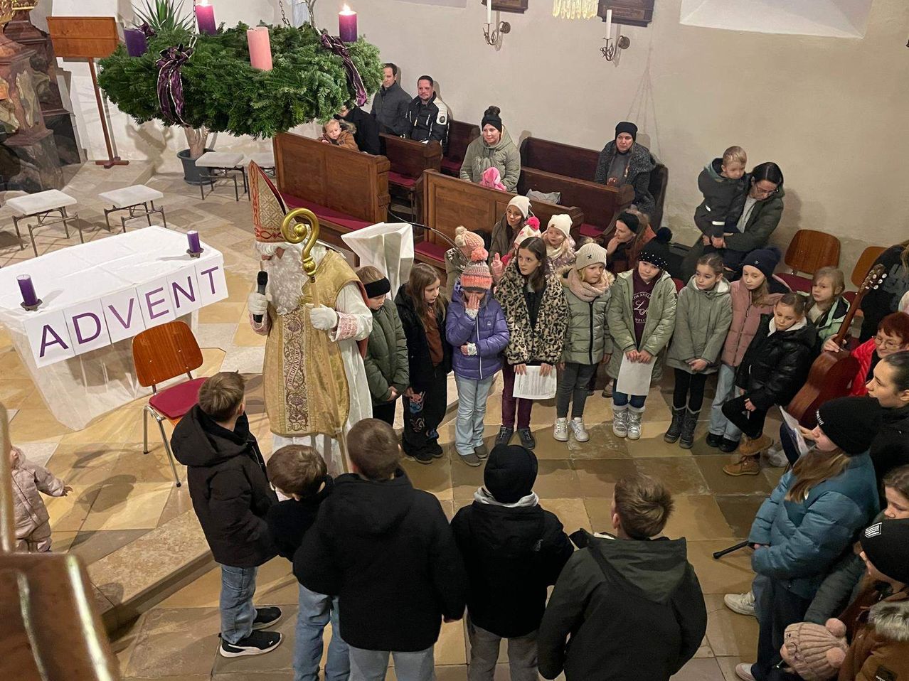 In einer Kirche spricht ein Priester zu einem Kreis von Kindern, von denen einige Papiere halten, während andere aufmerksam zuhören. Ein Banner mit dem Wort 'VENT' ist sichtbar. Erwachsene stehen in der Nähe, einige beobachten die Szene.