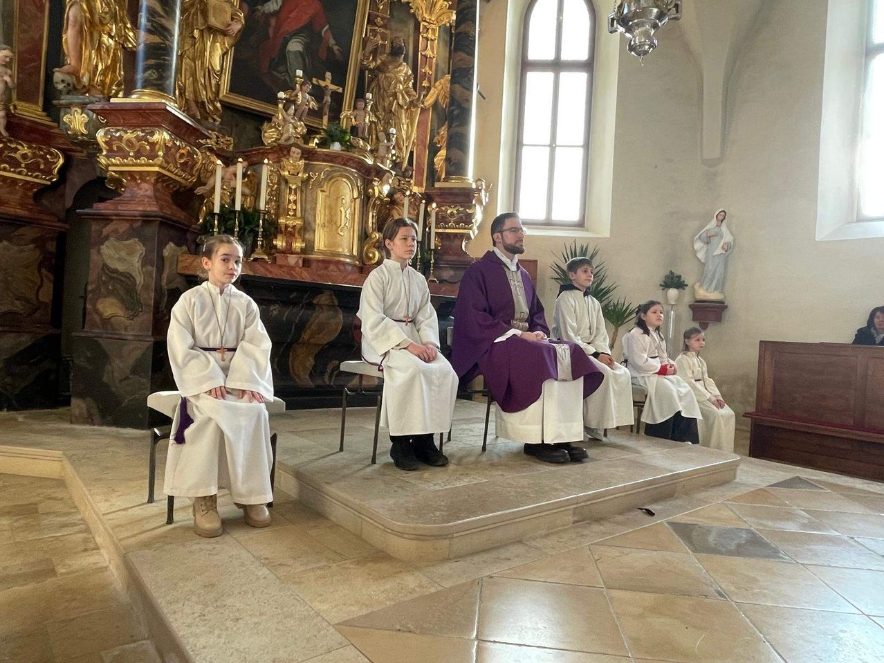 Ein Priester und mehrere Kinder in weißen Gewändern sitzen auf Stühlen in einer Kirche und blicken nach vorne. Dahinter ist ein Altar mit Kerzen und Statuen zu sehen.