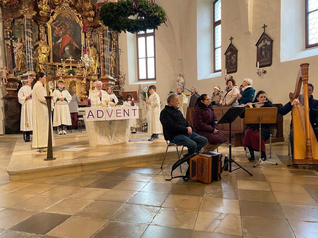 Ein Adventsgottesdienst findet in einer Kirche statt. Geistliche und Teilnehmer sind anwesend. Ein Banner mit der Aufschrift 'ADVENT' ist prominent angebracht. Musikinstrumente und Notenblätter sind aufgestellt.