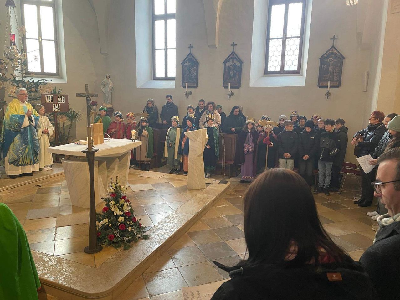Eine Gruppe von Menschen in Kostümen versammelt sich um einen Altar in einer Kirche. Der Altar hat ein Kreuz und Blumen, und es gibt Statuen und Gemälde an den Wänden.
