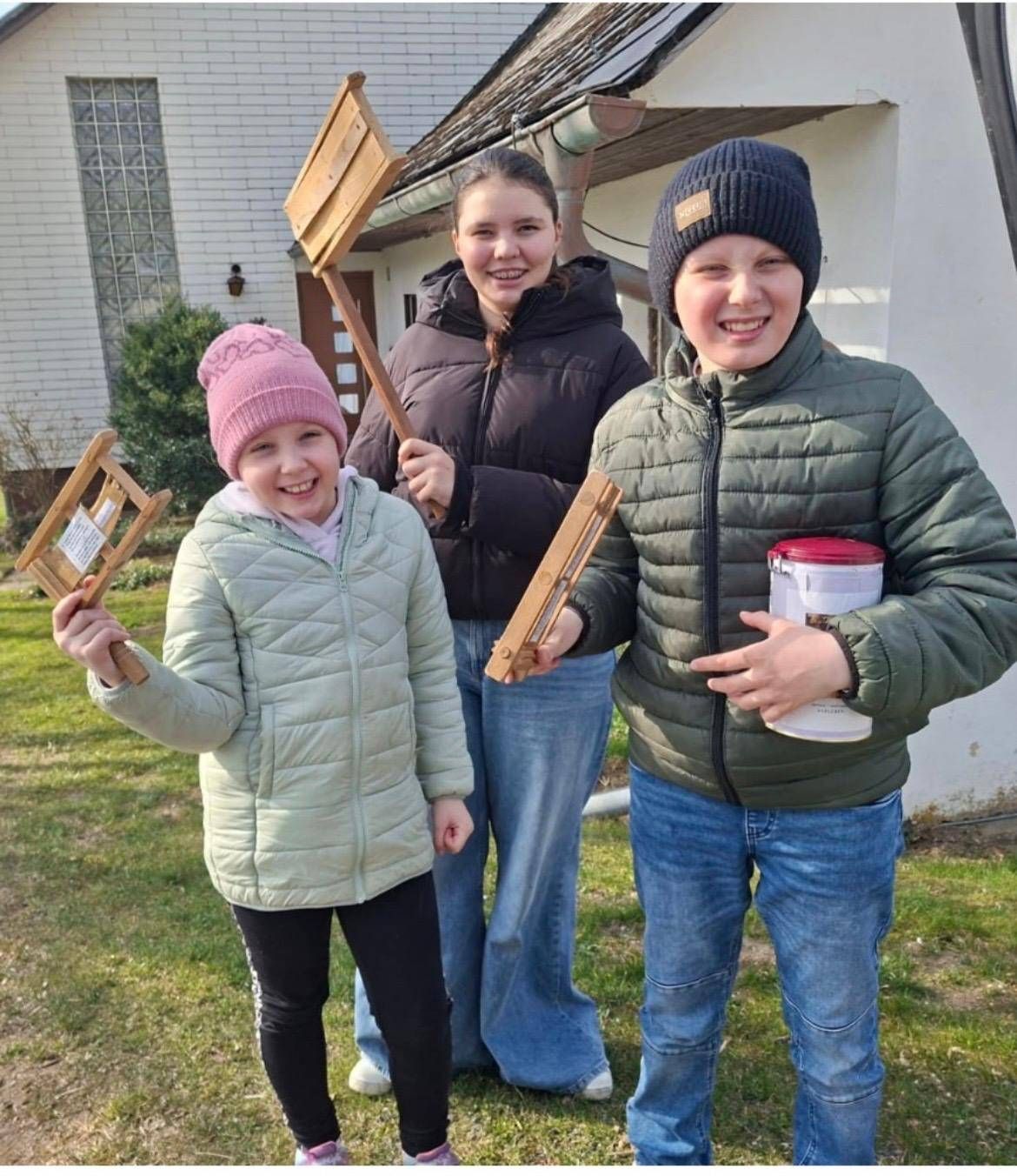 Three children in winter clothing stand outside a house. Two hold wooden objects, one holds a container.