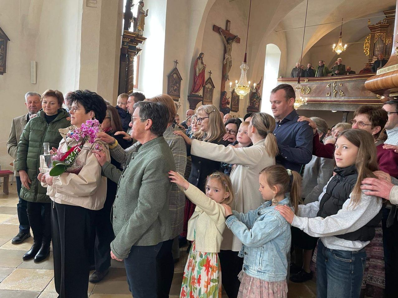 A group of people gather in a church, some holding flowers, with religious symbols and chandeliers in the background.