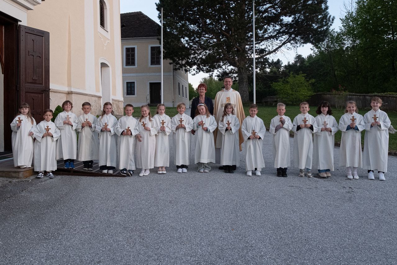 A group of children in white robes and a priest in a golden robe stand outside in front of a church.