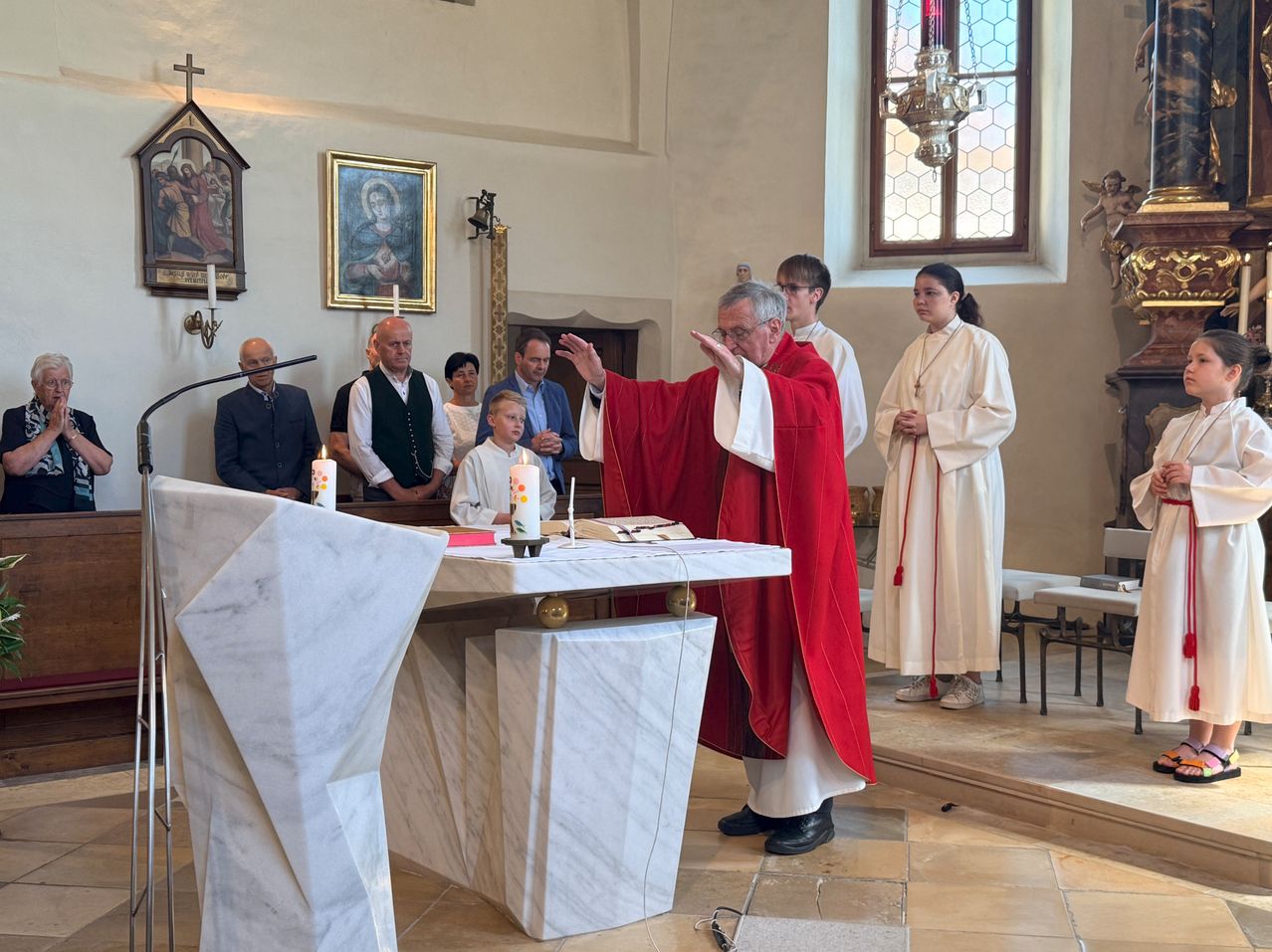 Ein Priester leitet eine kirchliche Zeremonie. Ein Mann in einer roten Robe steht am Altar und deutet. Kerzen und ein Buch liegen auf dem Altar.