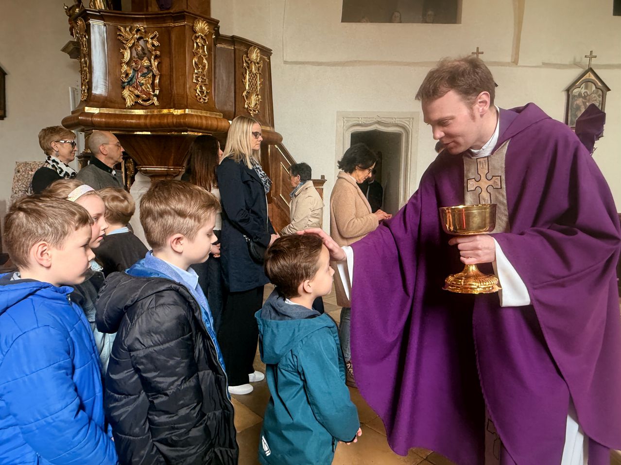 Ein Priester in einer violetten Robe segnet einen jungen Jungen, der einen goldenen Kelch hält, während andere in einer Kirche zusehen.