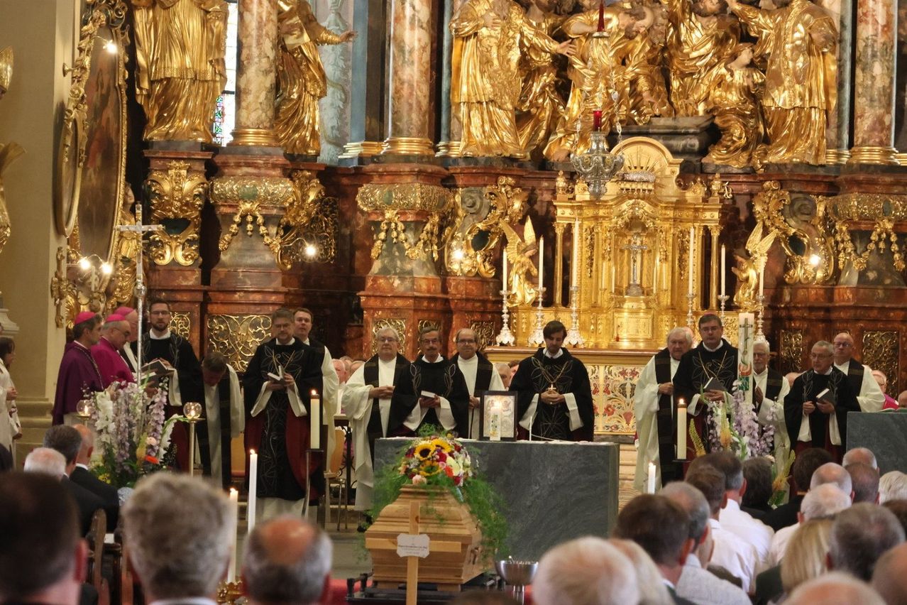 Eine Gruppe von Priestern steht vor einem Altar in einer Kirche mit Kerzen und Blumen. Ein Sarg ist auf dem Altar platziert.
