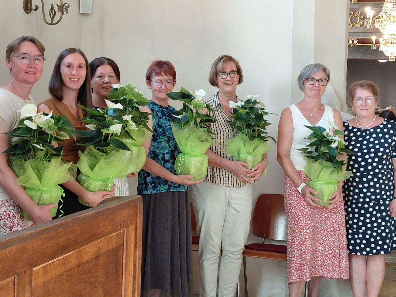 Fünf Frauen stehen zusammen und halten jeweils einen Strauß weißer Blumen, die in grünes Seidenpapier eingewickelt sind, und lächeln für ein Foto in einem Raum mit einer weißen Wand und einem Holzstuhl.