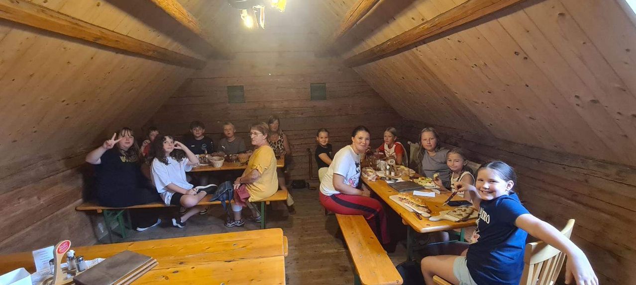 Eine Gruppe von Menschen sitzt an Holztischen in einem hölzernen Raum, möglicherweise einem Restaurant, mit Essen und Getränken auf den Tischen.