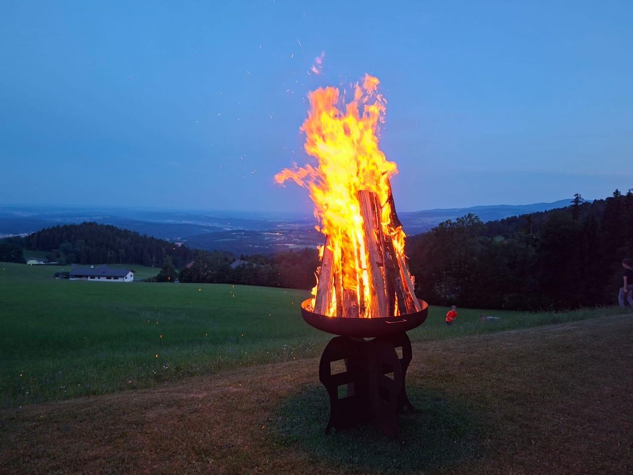 Ein großes Lagerfeuer brennt hell in einem Feld bei Dämmerung, umgeben von Bäumen und einem entfernten Haus. Die Flammen flackern und knistern und werfen ein warmes Licht.
