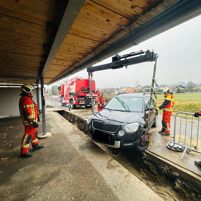 Rettungskräfte benutzen einen Kran, um einen schwarzen SUV aus einer Rinne unter einem hölzernen Überhang zu entfernen. Ein Feuerwehrwagen ist in der Nähe geparkt.