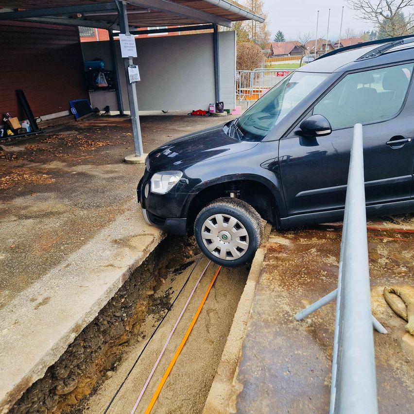 Ein schwarzer SUV ist an einem Bordstein neben einer Garage mit Carport geparkt. In der Nähe befindet sich eine Metallbarriere und der Boden ist teilweise mit Erde bedeckt.
