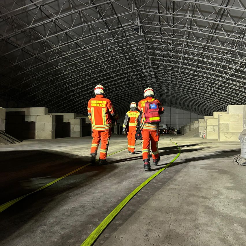 Three firefighters in orange uniforms walk through a tunnel with a metal roof and a yellow line on the floor. They are equipped with helmets and gloves.