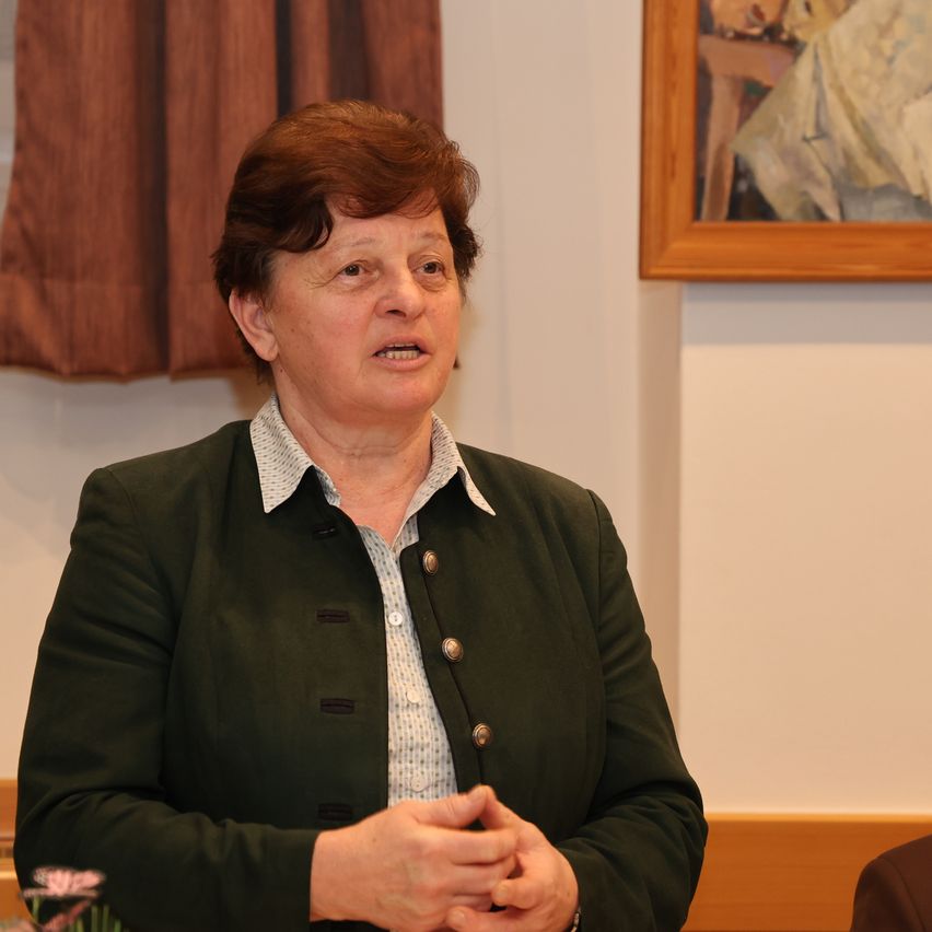 Eine ältere Frau mit roten Haaren steht und spricht in einem Raum mit weißer Wand und braunem Vorhang. Sie trägt einen grünen Blazer.