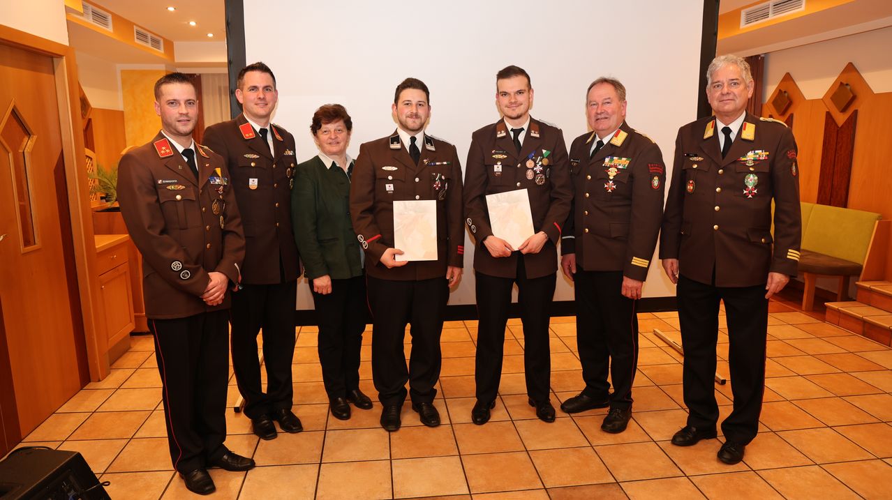 Eine Gruppe von Militäroffizieren in braunen Uniformen steht in einer Reihe und hält Urkunden, wobei eine Frau in einer grünen Jacke hinter ihnen steht.