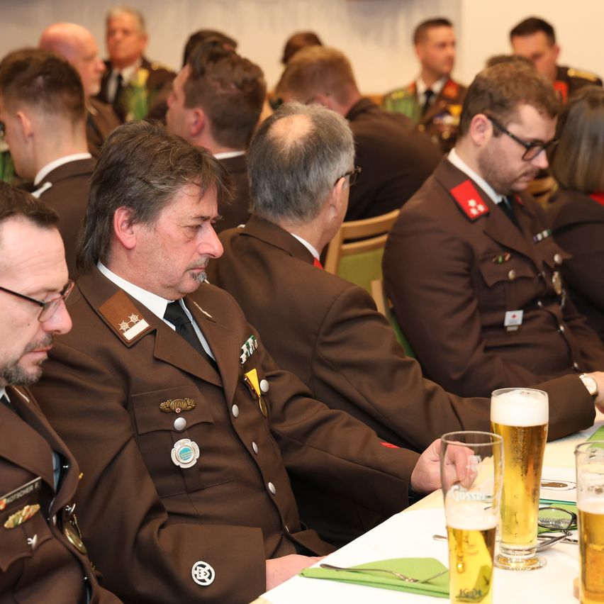 Eine Gruppe uniformierter Männer in einer Besprechung, in Stühlen sitzend, zwei mit Brille, einer mit einem Glas Bier und andere beobachten.