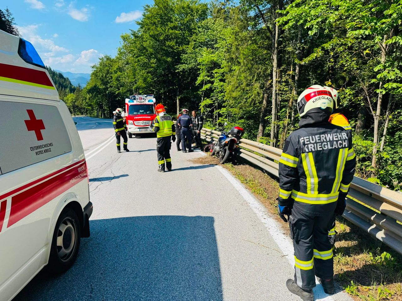 Rettungskräfte in Schutzkleidung stehen am Straßenrand, in der Nähe eines Motorradunfalls. Ein Feuerwehrwagen ist hinter ihnen geparkt.