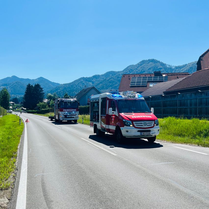 Zwei rote und weiße Feuerwehrfahrzeuge fahren auf einer Straße, mit Bergen und Häusern im Hintergrund.