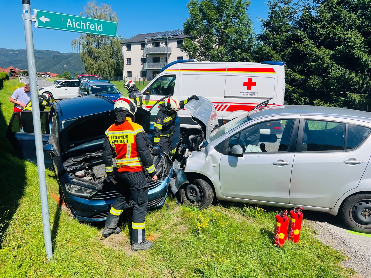 Rettungskräfte in Schutzausrüstung inspizieren ein beschädigtes Fahrzeug. Ein Krankenwagen mit rotem Kreuz ist in der Nähe, und zwei Feuerlöscher stehen auf dem Gras.