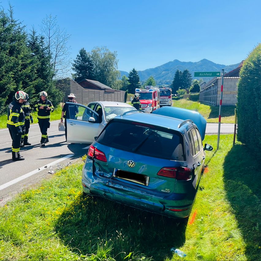 Rettungskräfte untersuchen einen Autounfall auf einer Straße. Ein blaues Auto ist auf dem Gras neben der Straße. Eine Person sitzt am Steuer.