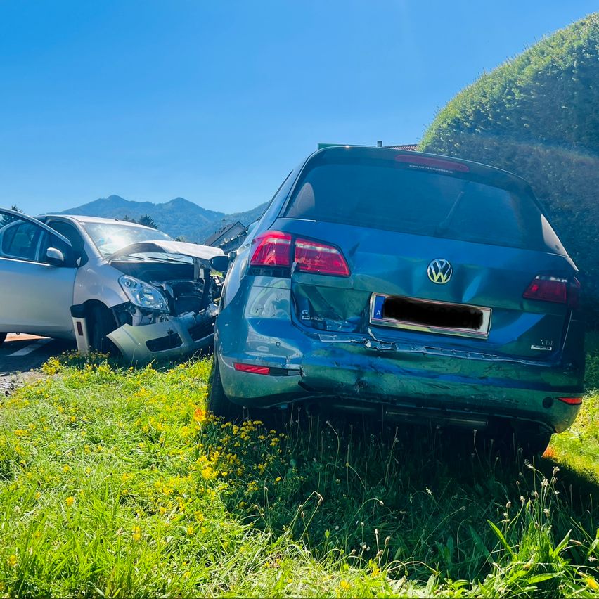 Zwei Autos auf einem Grasbereich geparkt, eines mit beschädigter Stoßstange und das andere mit beschädigter Front. Der Himmel ist klar und Berge sind in der Ferne sichtbar.