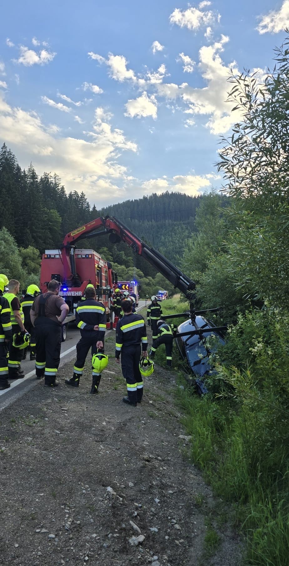 Feuerwehrleute stehen neben einem umgekippten Fahrzeug, das von einem Kran angehoben wird. Ein Wald und eine bewölkte Sonne sind im Hintergrund.