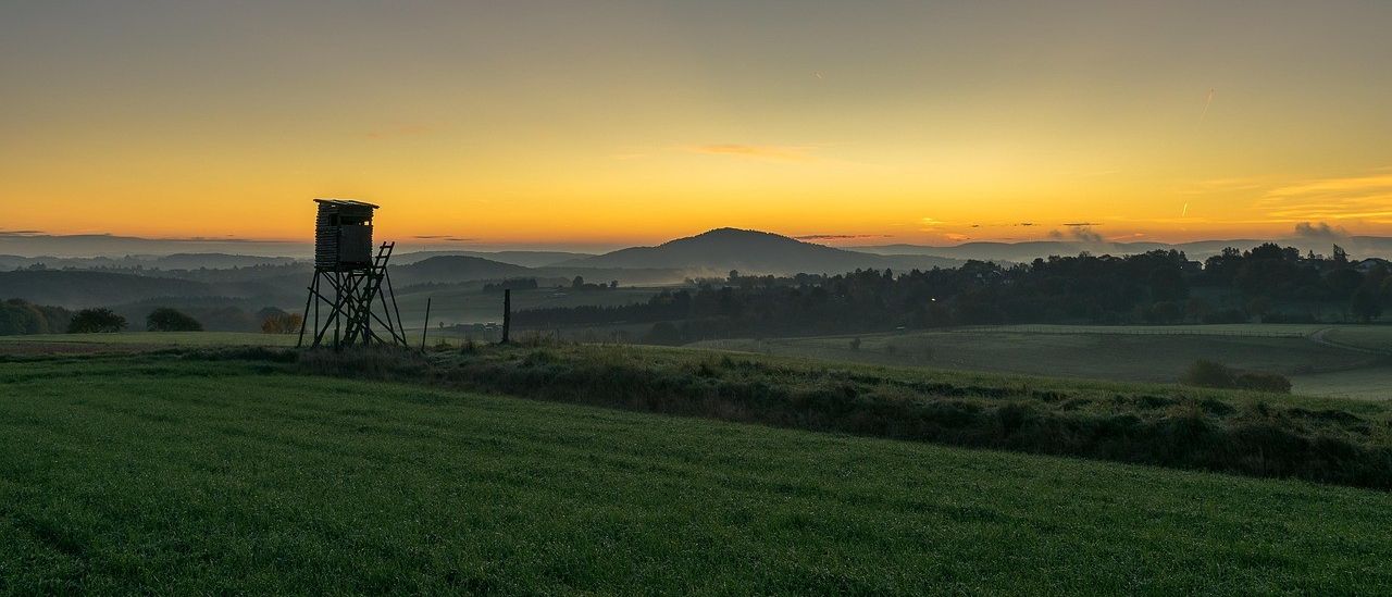 Bei Sonnenuntergang steht ein Jagdturm in einem grünen Feld. Berge und Hügel sind in der Ferne. Der Himmel ist orange und pink.