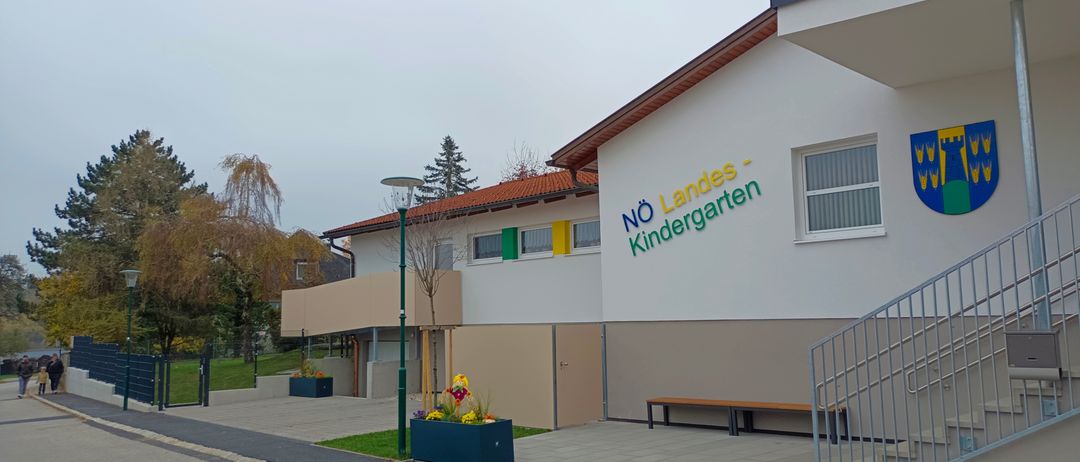 Ein Kindergartengebäude mit einem roten Dach und einem Schild, das 'No Landes-Kindergarten' steht. Das Gebäude hat eine Bank und ein Metallgeländer. Es gibt Bäume und eine Straßenlaterne in der Gegend.