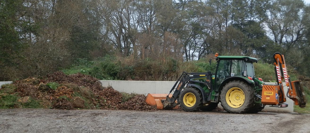 Ein grüner Traktor mit einem gelben Frontlader steht auf einer Kiesfläche, mit einem Haufen Blätter in der Nähe.