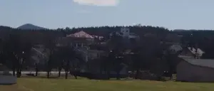 Eine Landschaft mit einem breiten grünen Feld und mehreren Gebäuden und Bäumen. Der Himmel ist blau mit einer weißen Wolke in der Ferne.