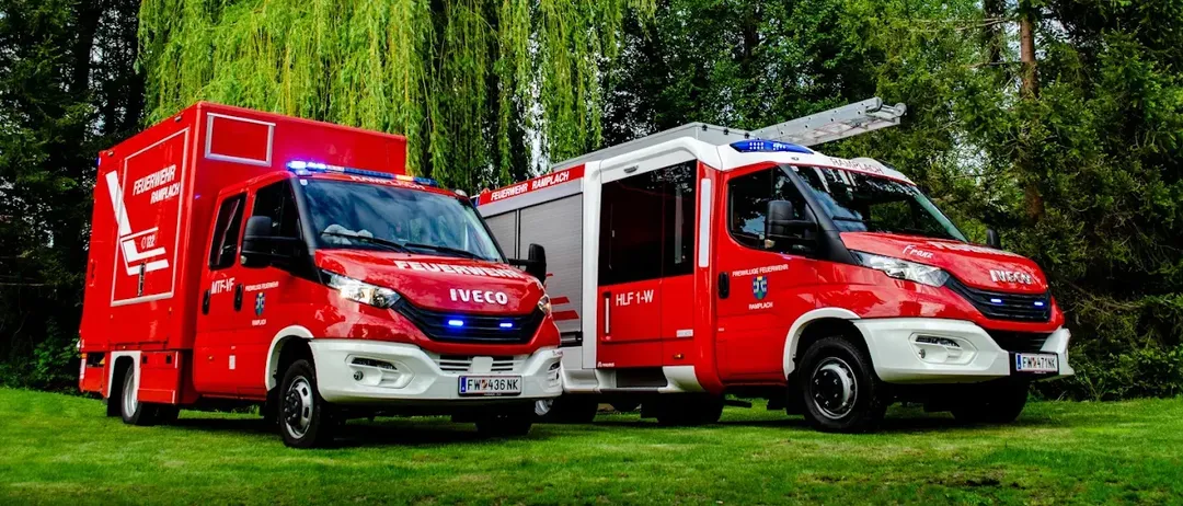 Zwei rote Notfallfahrzeuge, eines mit dem Wort Iveco auf der Front und das andere mit HLF 1-W an der Seite, geparkt auf einem Rasenfeld mit Bäumen im Hintergrund.