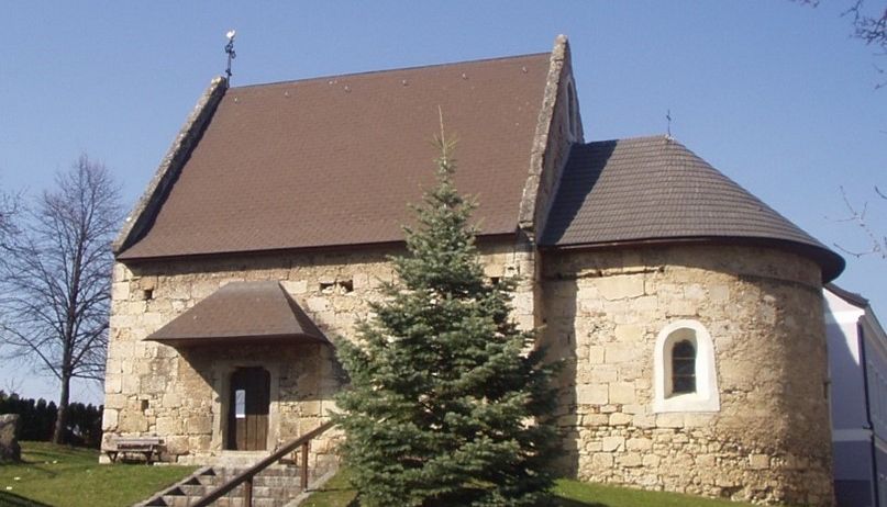 Eine alte Steinkirche mit einem braunen Dach und einer kleinen Tanne vor ihr.