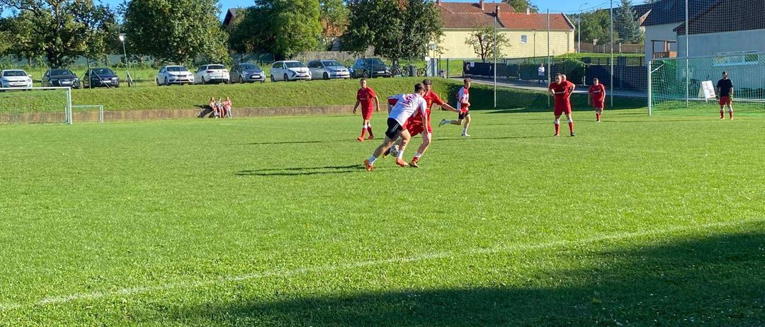 Eine Gruppe von Fußballspielern in roten Trikots spielt auf einem Feld mit einem Tor in der Ferne. Bäume und Gebäude sind im Hintergrund zu sehen.