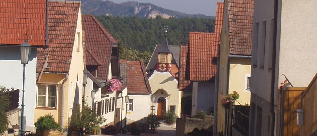 Eine schmale Straße gesäumt von traditionellen Häusern mit roten Dächern und einer kleinen Kirche in der Ferne. Der Himmel ist klar und blau.
