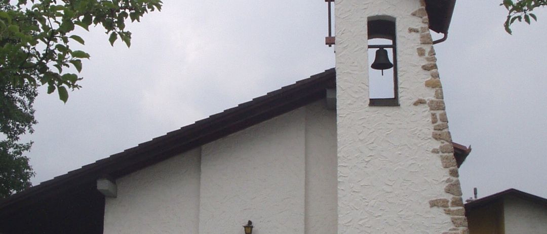 Eine kleine weiße Kirche mit einer braunen Tür und einem Glockenturm, umgeben von einem Feld und Bäumen, unter einem bewölkten Himmel.