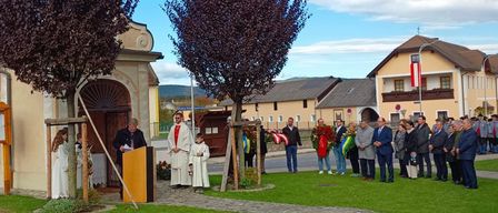 Eine Gruppe von Menschen versammelt sich vor einer Kirche, wobei zwei Priester an einem Podium stehen. Die Umgebung umfasst Bäume, einen klaren blauen Himmel und eine kleine Menschenmenge.